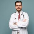 Smiling medical worker in white coat and tie. A portrait of a doctor posing against gray background
