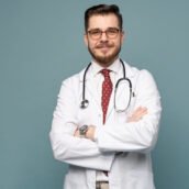 Smiling medical worker in white coat and tie. A portrait of a doctor posing against gray background